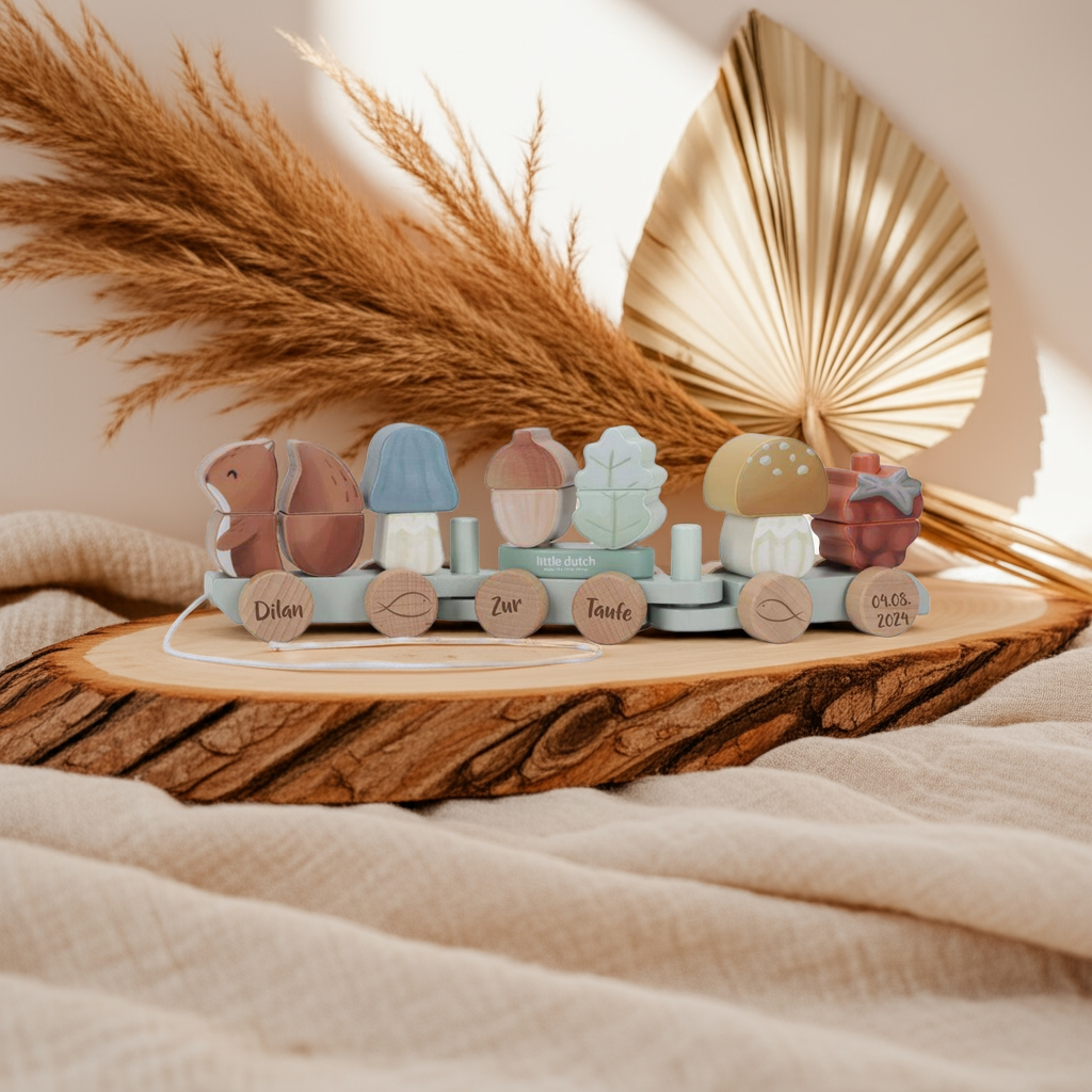 The Holz-Zug Eichhörnchen Forest Friends "Zur Taufe" by Little Dutch, a pastel wooden pull toy with nature-themed blocks, sits on a wood tray among beige fabric and pampas grass, softly lit with a palm leaf in the background.